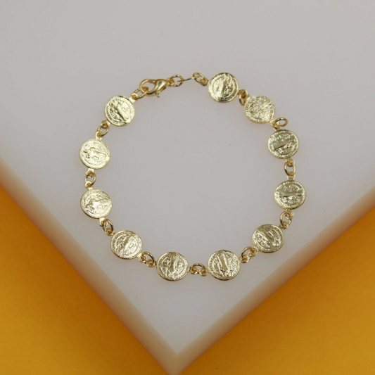 A delicate gold Saint Benedict coin charm bracelet rests on a white and yellow display surface.