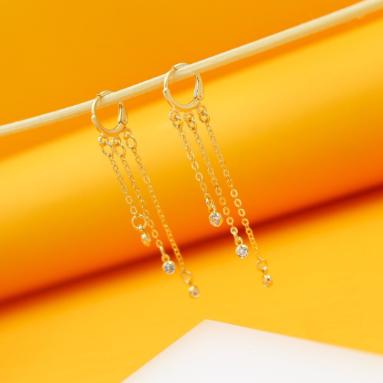 Two dangling gold huggie hoop earrings with delicate chain fringes and sparkling cubic zirconia stones hang from a wooden stick against a bright yellow background.