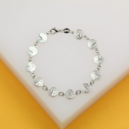 A silver Saint Benedict medal charm bracelet rests on a white and yellow display block.