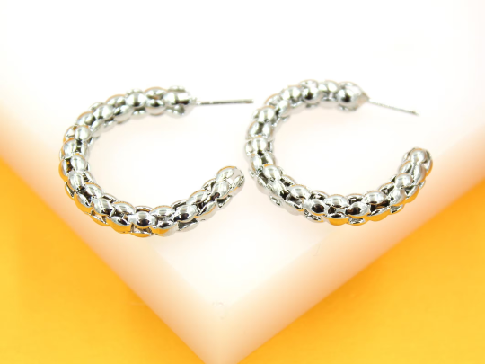 A pair of shiny silver textured hoop earrings rests on a white display stand against a bright yellow background.