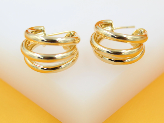 A pair of polished gold multi-layered hoop earrings sits neatly on a white and yellow display surface.