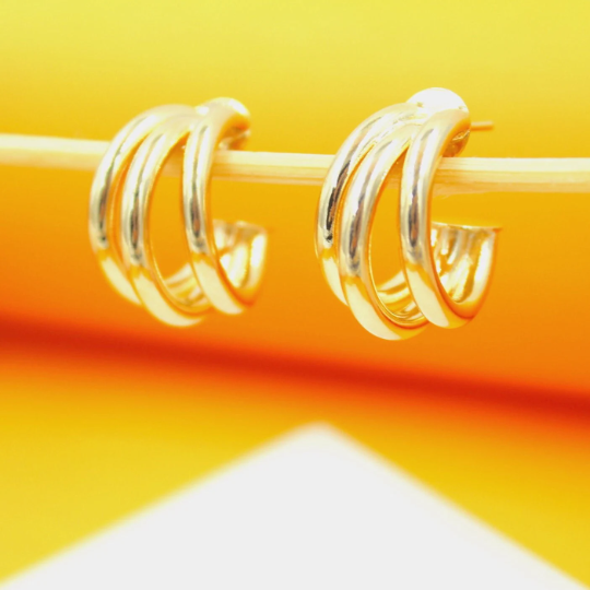 A pair of polished gold multi-hoop earrings rests gracefully on a wooden stick against a bright yellow and orange backdrop.