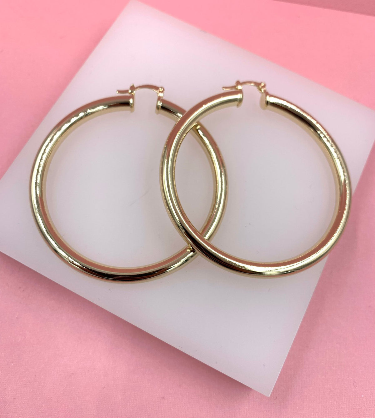A pair of polished gold hoop earrings rests on a white display block against a vibrant pink background.