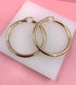 A pair of polished gold hoop earrings rests on a white display block against a vibrant pink background.