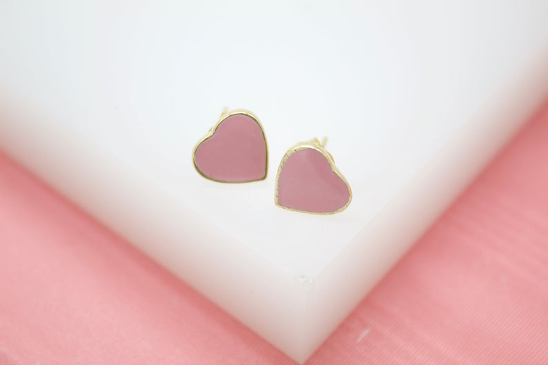 A pair of gold-rimmed pink heart stud earrings rest on a white display surface against a pink background.