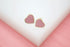 A pair of gold-rimmed pink heart stud earrings rest on a white display surface against a pink background.