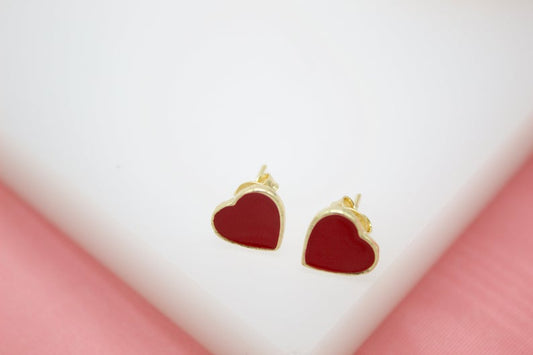 A pair of gold heart-shaped stud earrings featuring red enamel fill rest on a white display surface.
