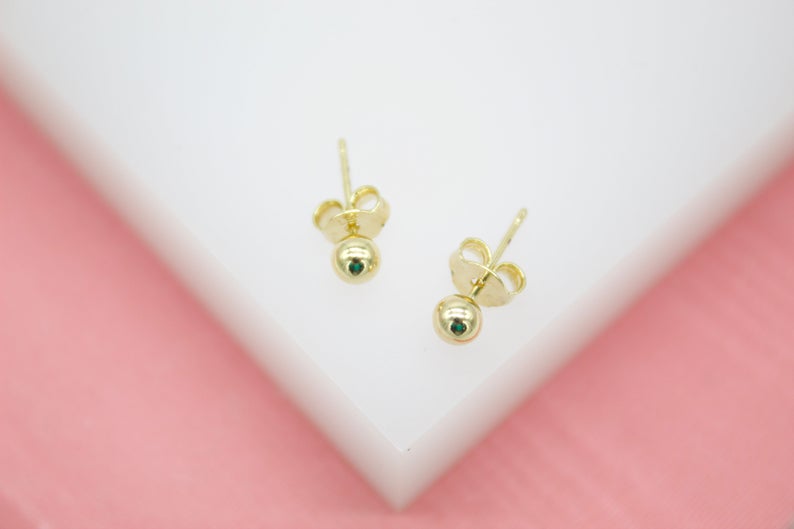 A pair of gold ball stud earrings featuring tiny emerald stones rests on a white display stand against a pink background.