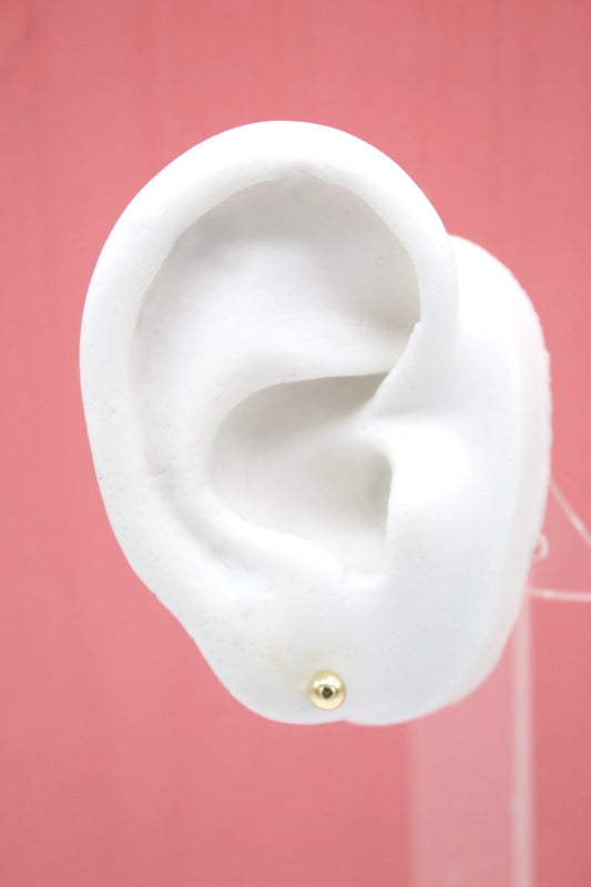 A small gold ball stud earring sparkles on a white mannequin ear against a solid pink background.