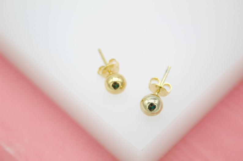 A pair of polished gold ball stud earrings rests against a white and pink background.