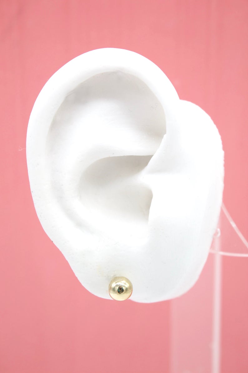 A white ear model displays a round gold ball stud earring on a pink background.