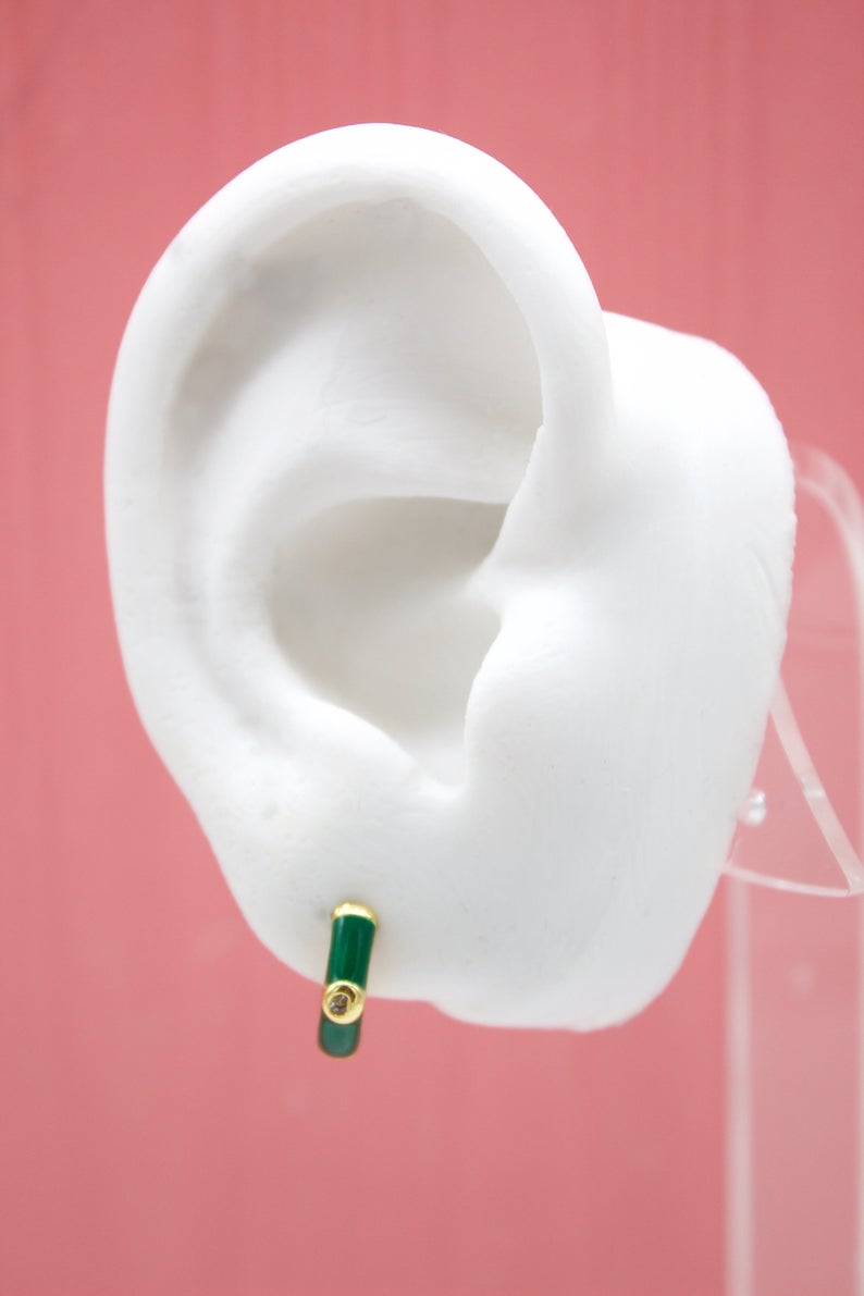 A white mannequin ear displays a gold huggie hoop earring featuring glossy green enamel and a small diamond accent.
