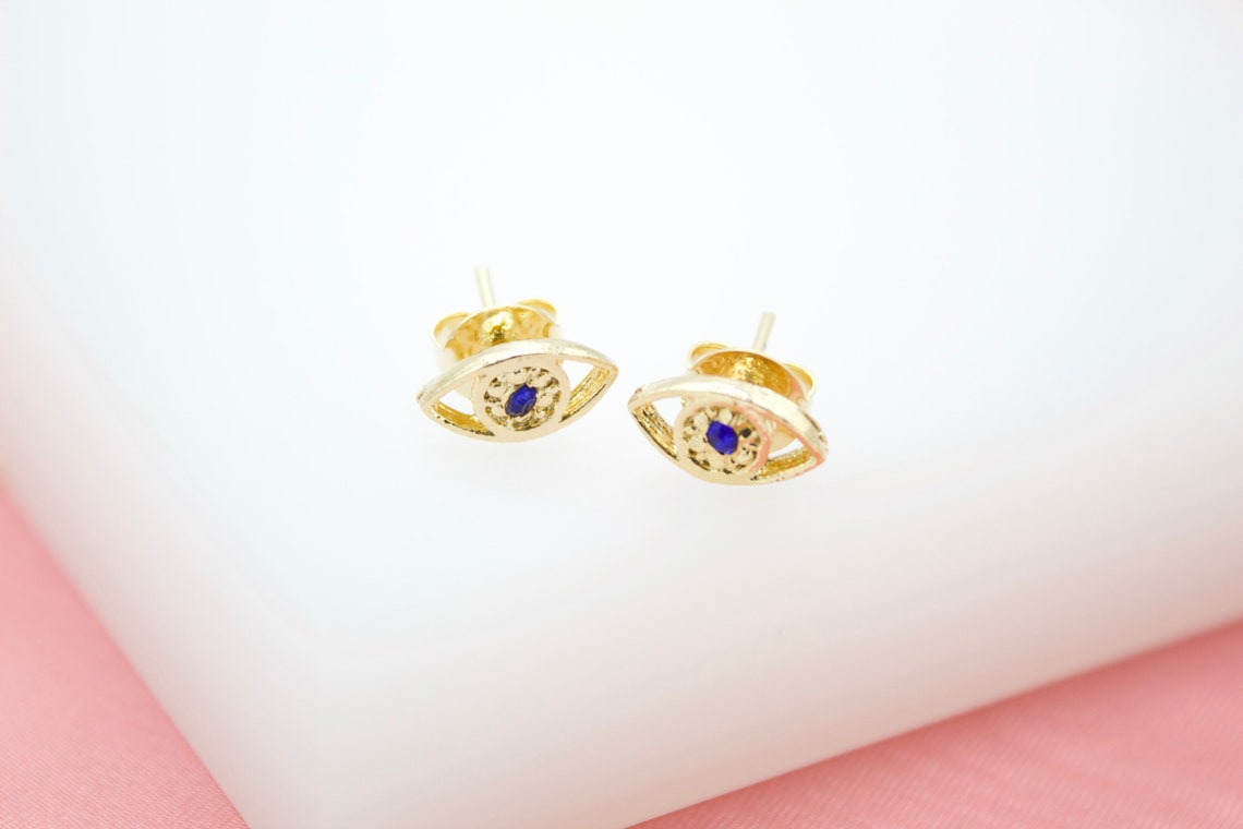 A pair of gold evil eye stud earrings featuring central blue stones rests against a white and pink background.