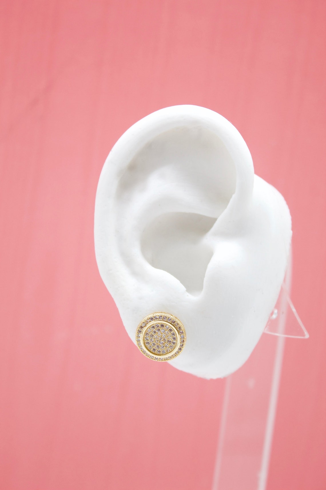 A white mannequin ear displays a round gold stud earring encrusted with sparkling pave diamonds against a solid pink backdrop.