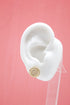 A white mannequin ear displays a round gold stud earring encrusted with sparkling pave diamonds against a solid pink backdrop.