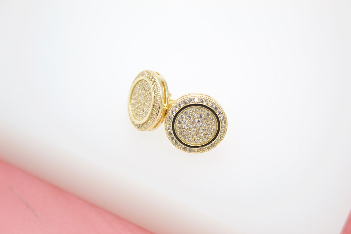 A pair of round gold stud earrings features intricate pavé-set CZ stones and a black enamel accent against a white background.