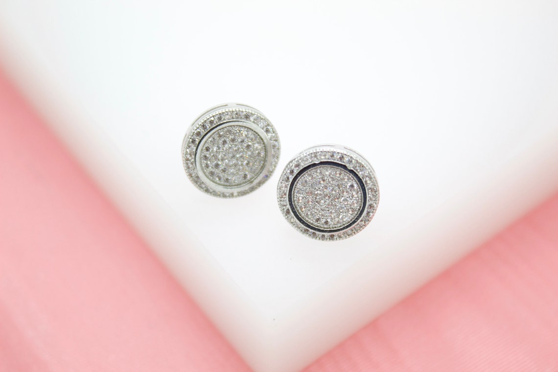 A pair of sparkling pavé diamond round stud earrings rests elegantly on a white surface against a soft pink background.