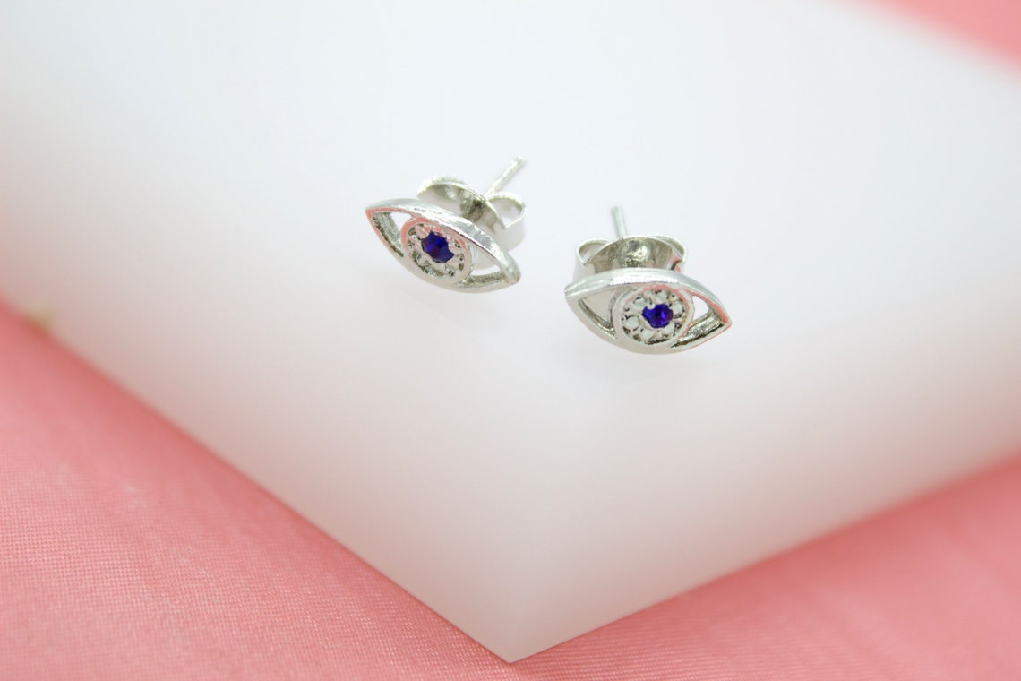 A pair of silver evil eye stud earrings featuring central blue crystals rest on a white display surface against a pink background.