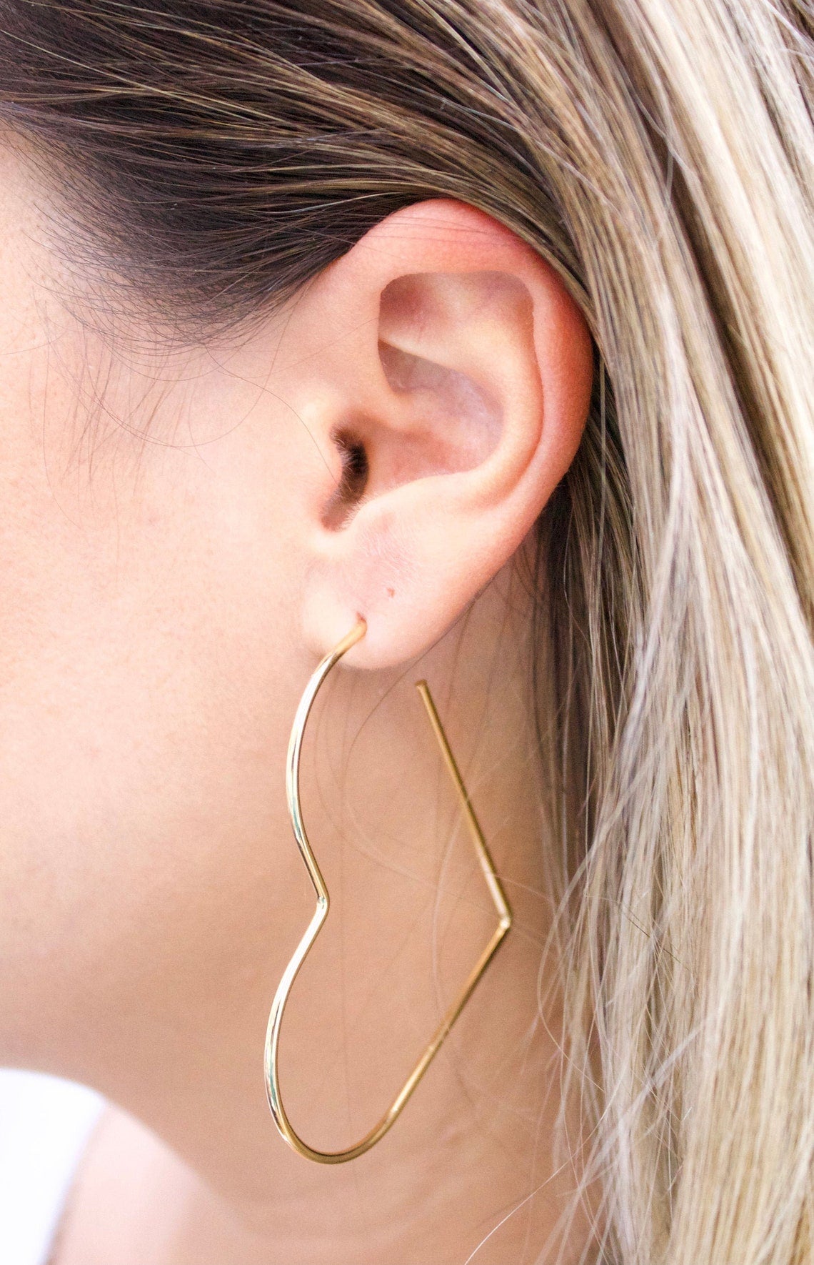 A woman with blonde hair wears a large, geometric gold heart-shaped hoop earring in her earlobe.