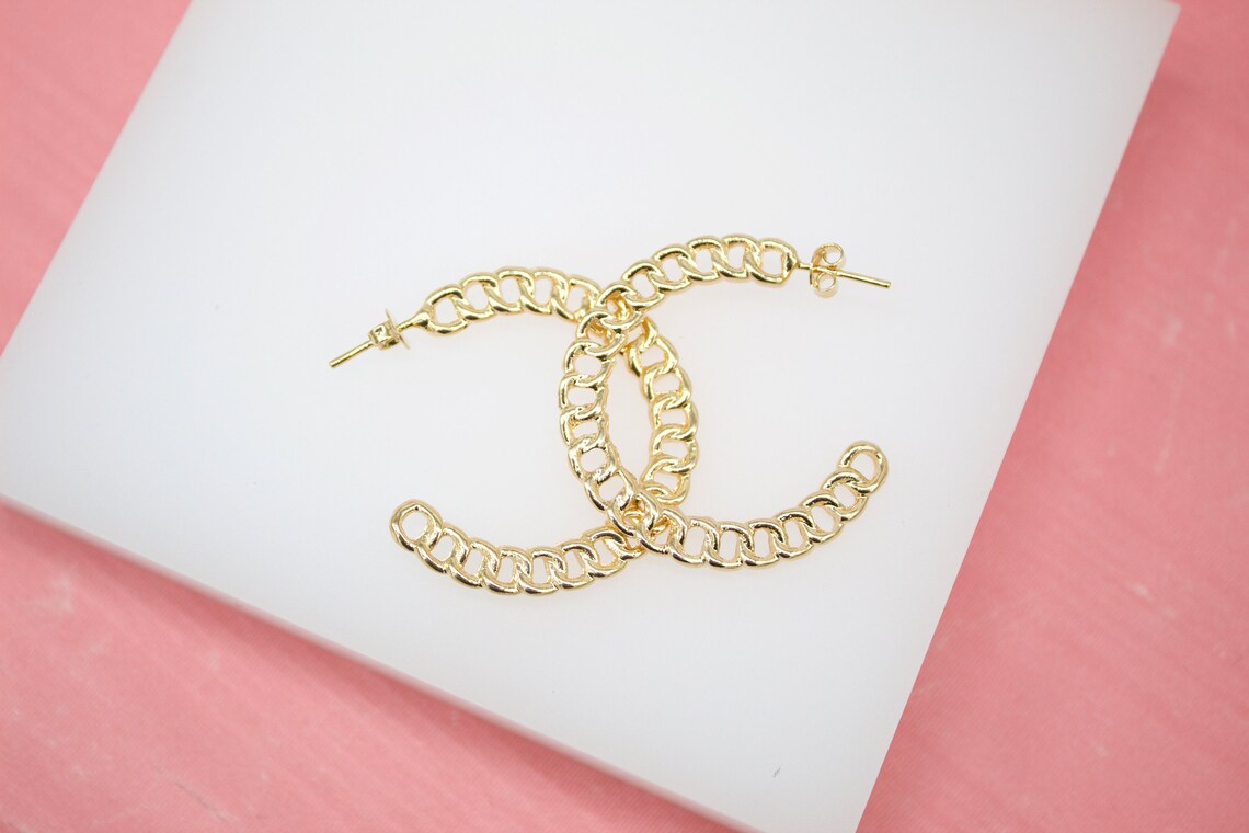 A pair of gold chain-link hoop earrings rests on a white display block against a pink background.