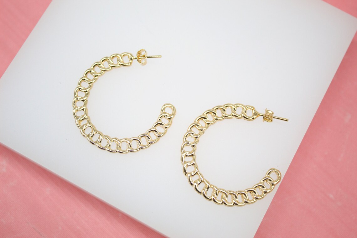 A pair of gold chain-link hoop earrings rests on a white display block against a soft pink background.