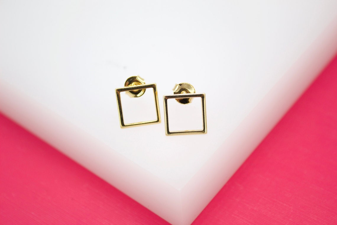A pair of minimalist gold square stud earrings rests on a white display surface against a bright pink background.