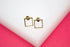 A pair of minimalist gold square stud earrings rests on a white display surface against a bright pink background.