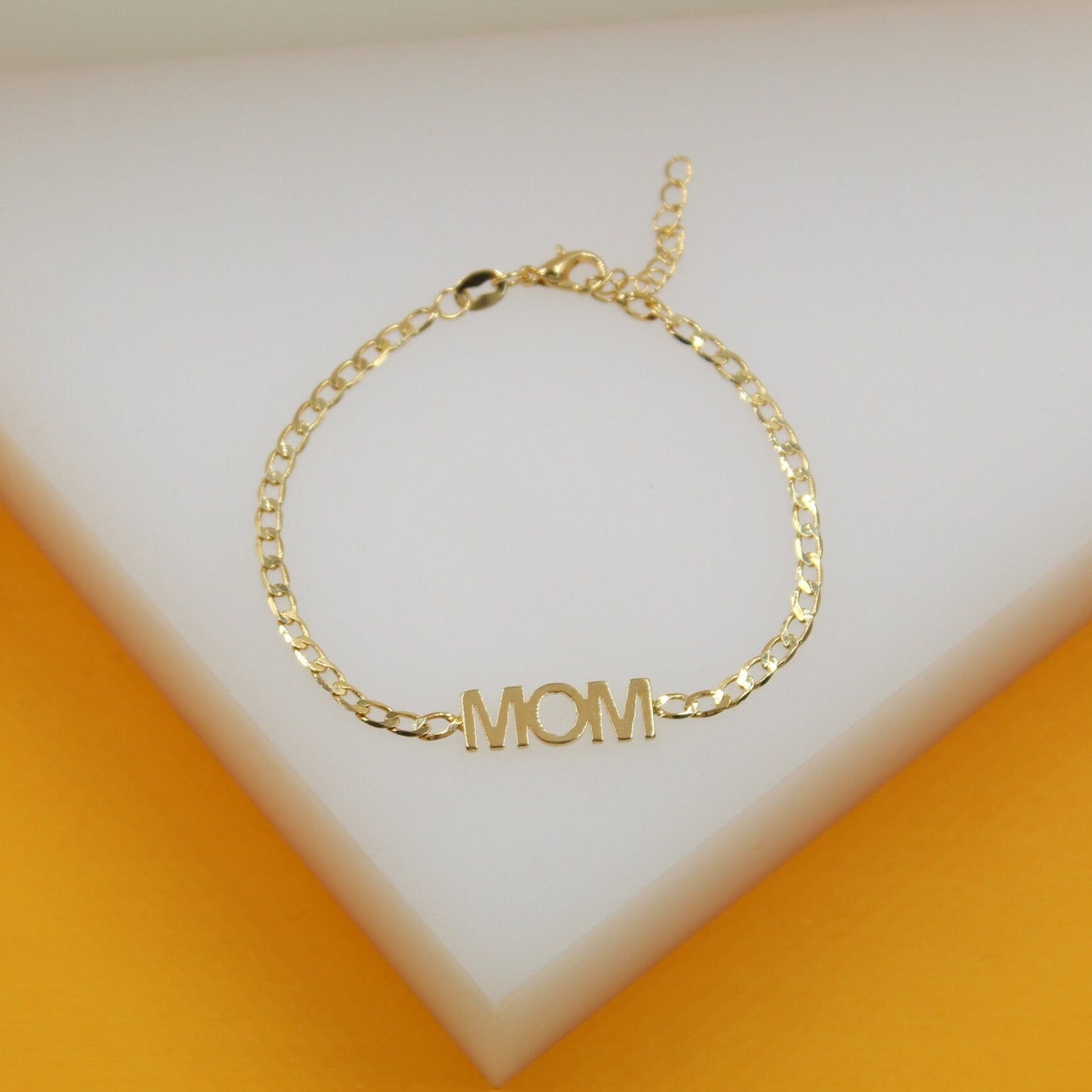 A delicate gold chain bracelet featuring the word "MOM" in block letters rests on a white display surface.