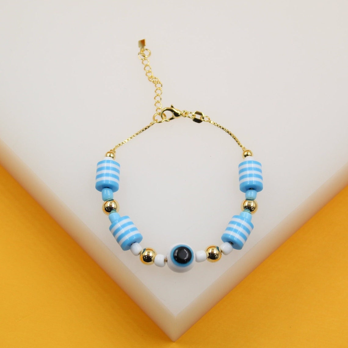 A delicate gold-chain bracelet features blue and white striped cylindrical beads and a central evil eye charm resting on a white display surface.