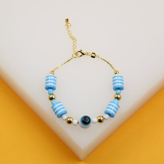 A delicate gold-chain bracelet features blue and white striped cylindrical beads and a central evil eye charm resting on a white display surface.