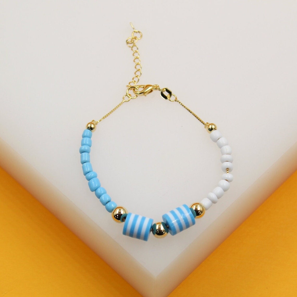 A dainty gold chain bracelet features alternating light blue and white seed beads accented by striped cylinder beads.
