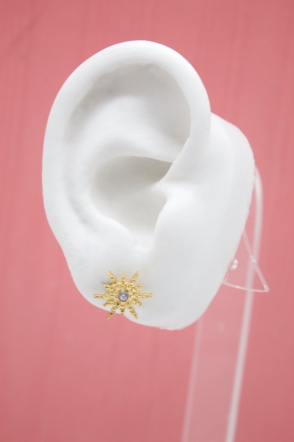 A white mannequin ear displays a gold starburst stud earring featuring a central gemstone.