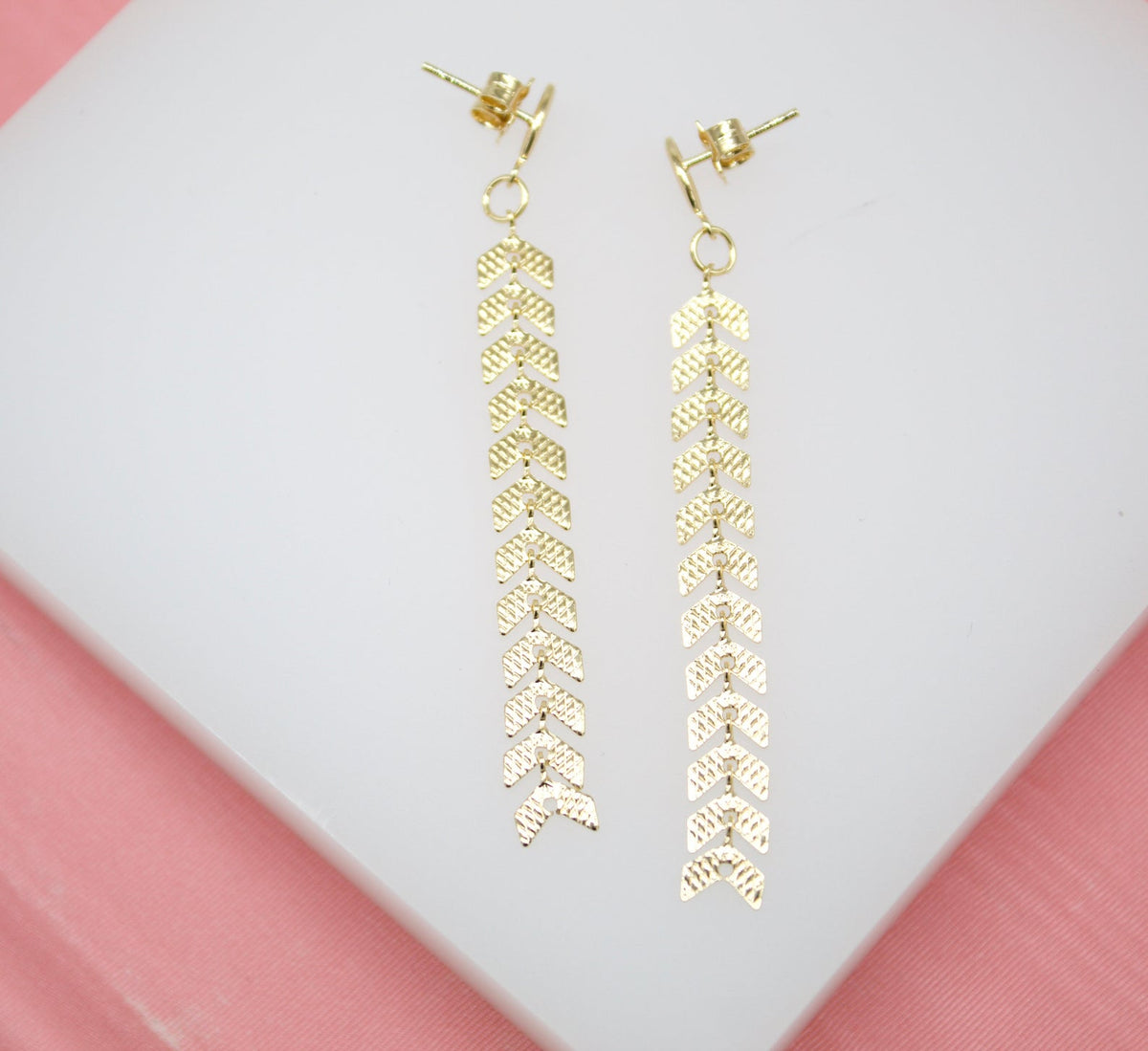 A pair of gold dangle earrings featuring long chains of textured chevron links rest on a white surface.