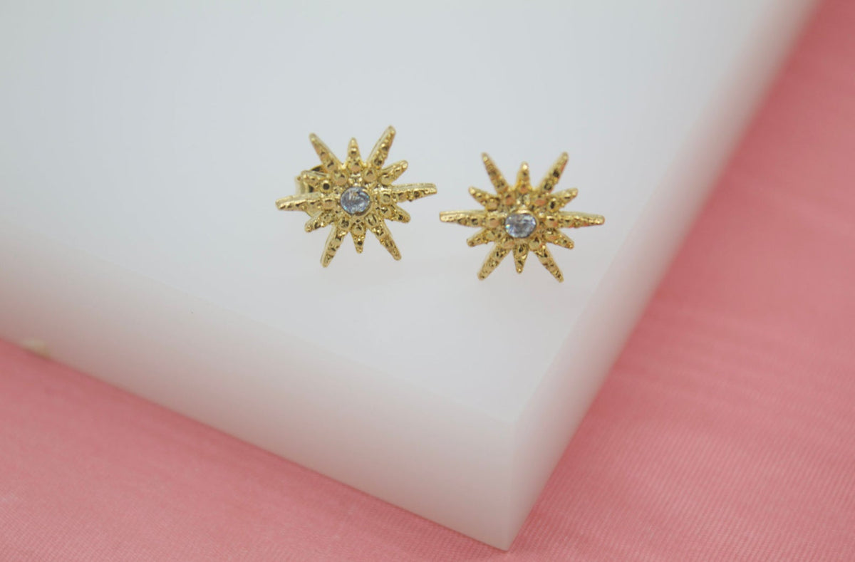 A pair of gold starburst stud earrings with central sparkling crystals rests on a white display block.