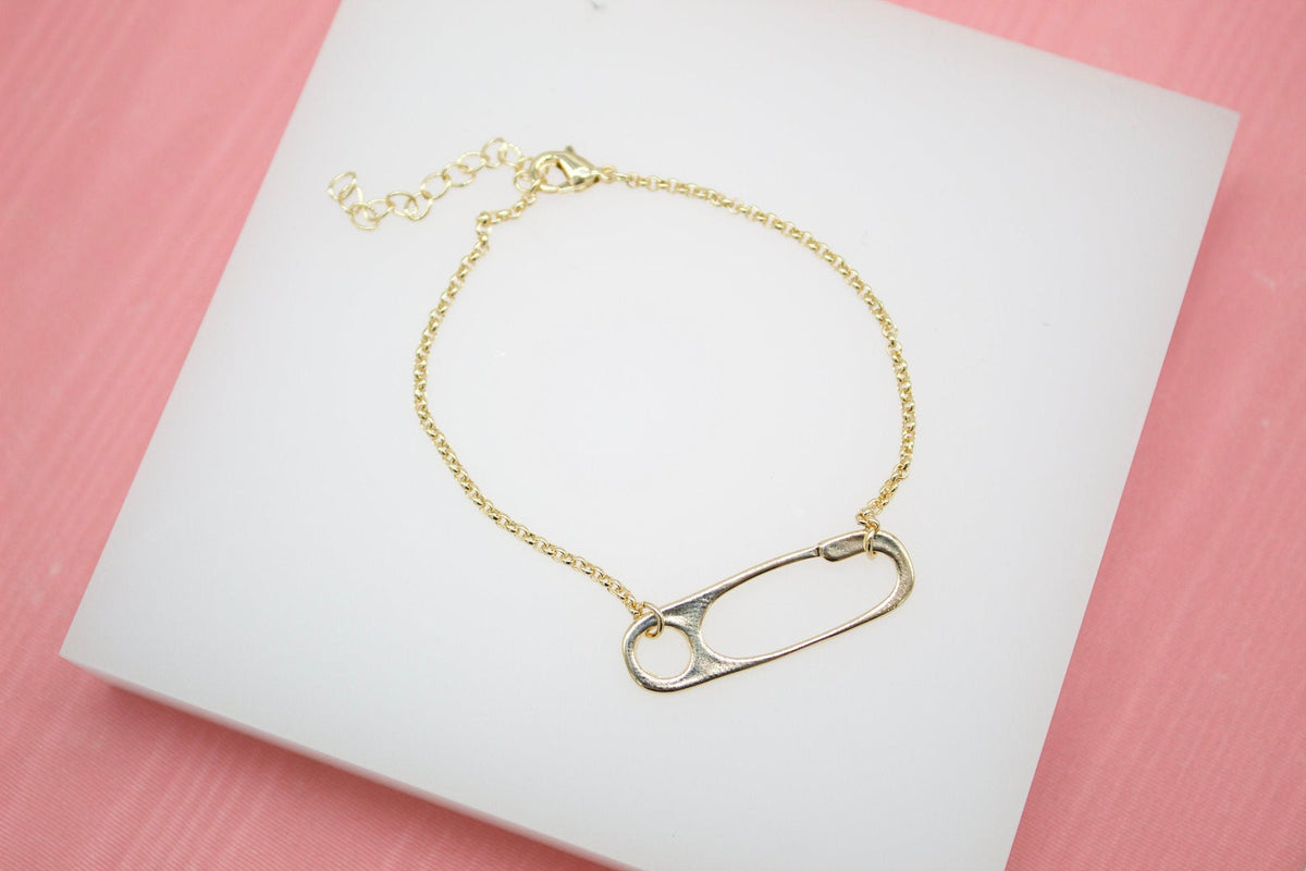 A delicate gold chain bracelet features a unique safety pin charm resting on a white display surface.