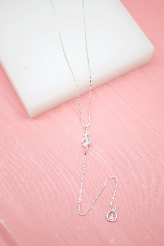 A delicate silver lariat necklace with sparkling cubic zirconia stones drapes gracefully against a pink and white backdrop.