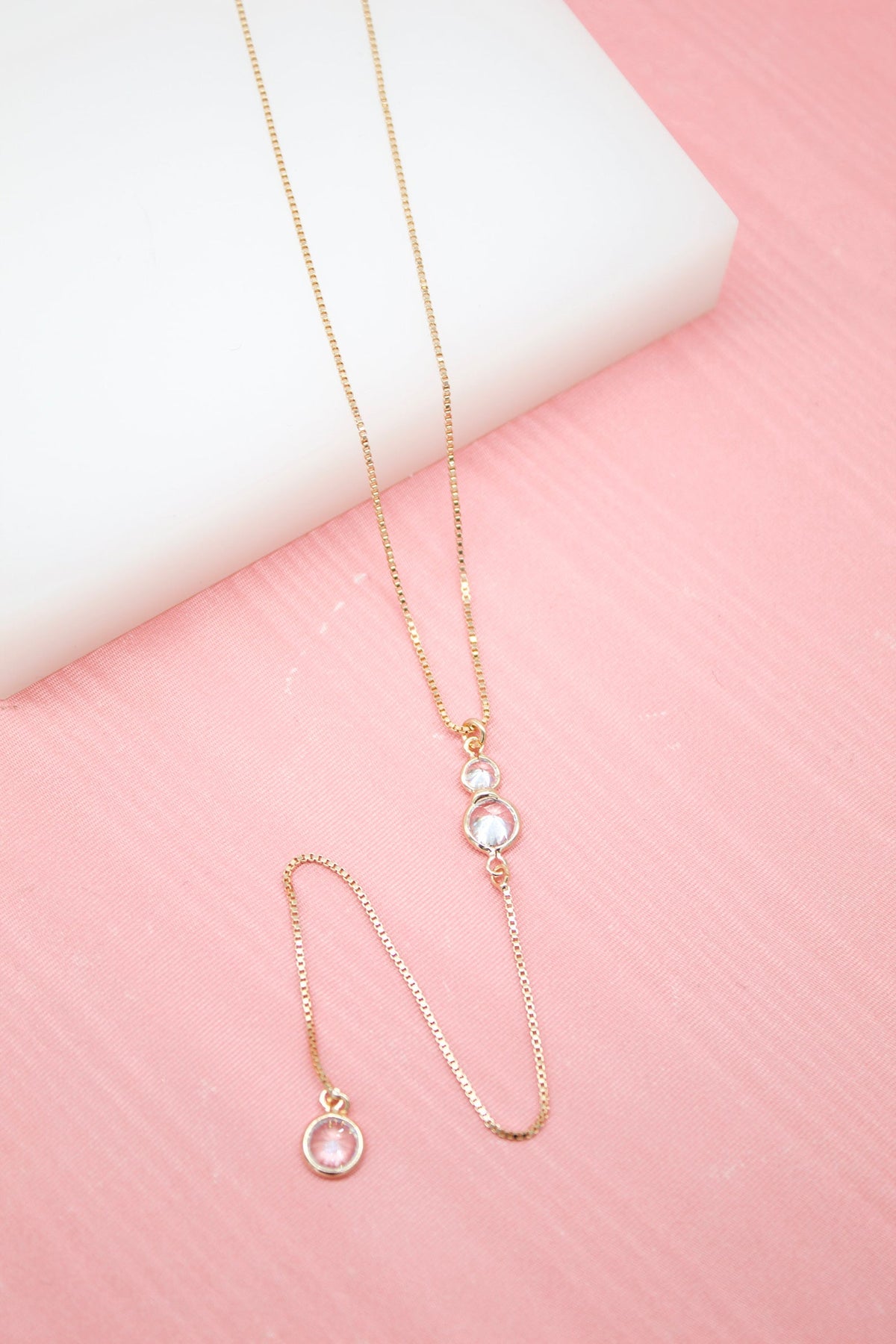 A delicate gold lariat necklace featuring dangling cubic zirconia crystals rests on a pink surface next to a white block.