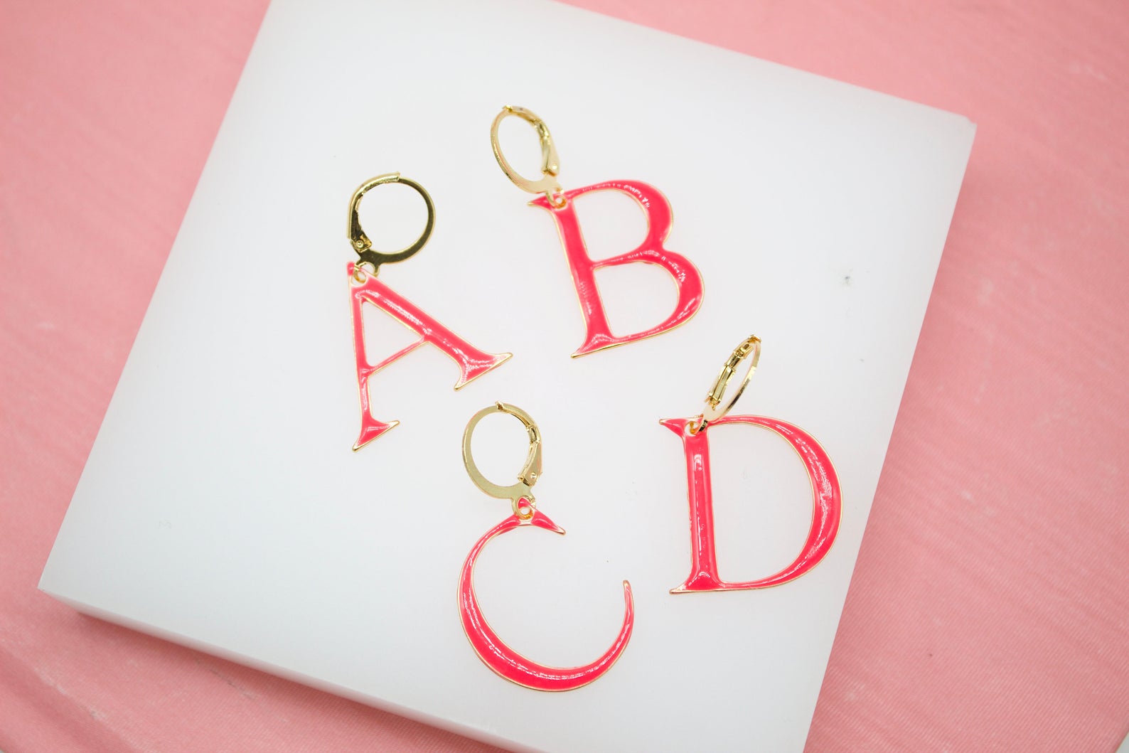 Four hot pink initial hoop earrings shaped like the letters A, B, C, and D rest on a white surface against a pink background.