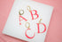 Four hot pink initial hoop earrings shaped like the letters A, B, C, and D rest on a white surface against a pink background.
