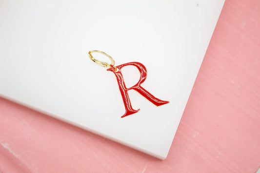 A gold-rimmed red initial R charm earring rests on a white display block.