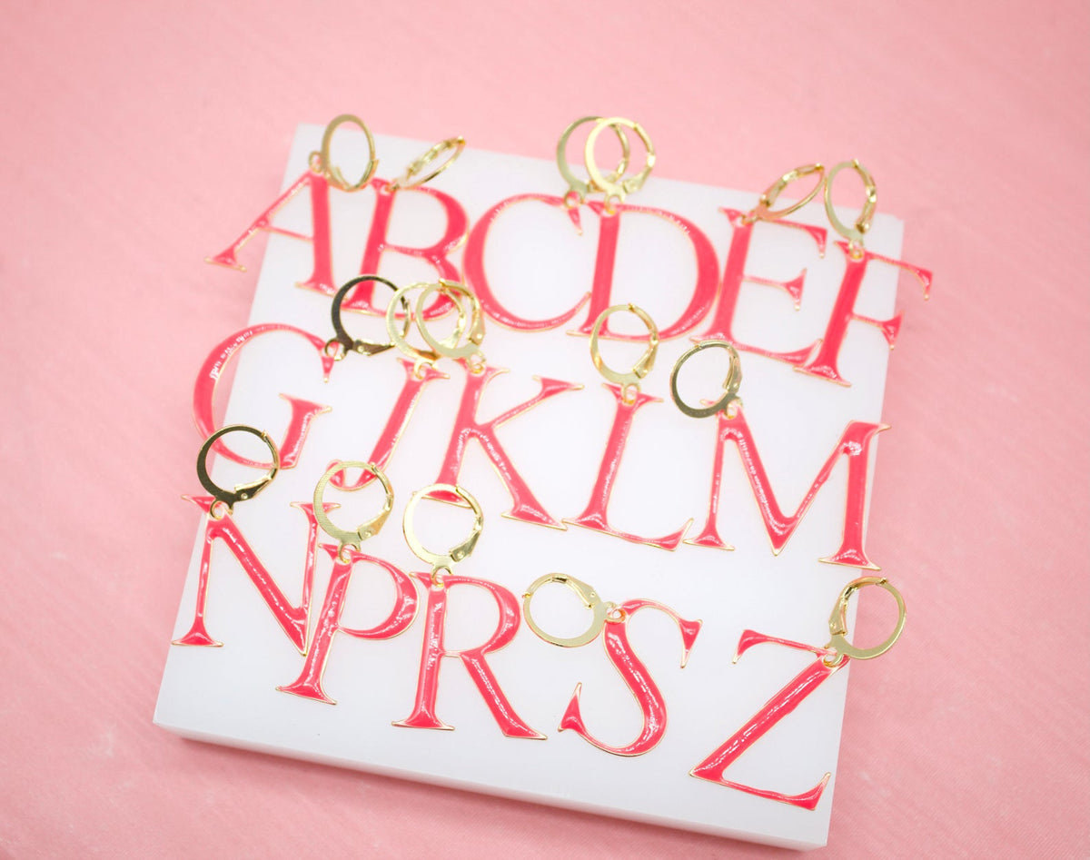 A collection of glossy hot pink enamel letter initial earrings with gold leverback clasps rests on a white display block against a soft pink background.