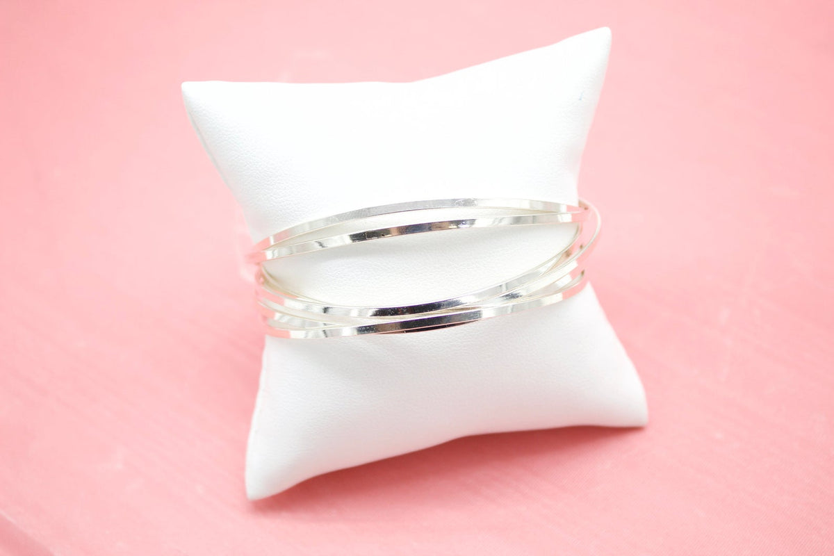 A stack of delicate silver bangles rests on a small white display pillow against a pink background.