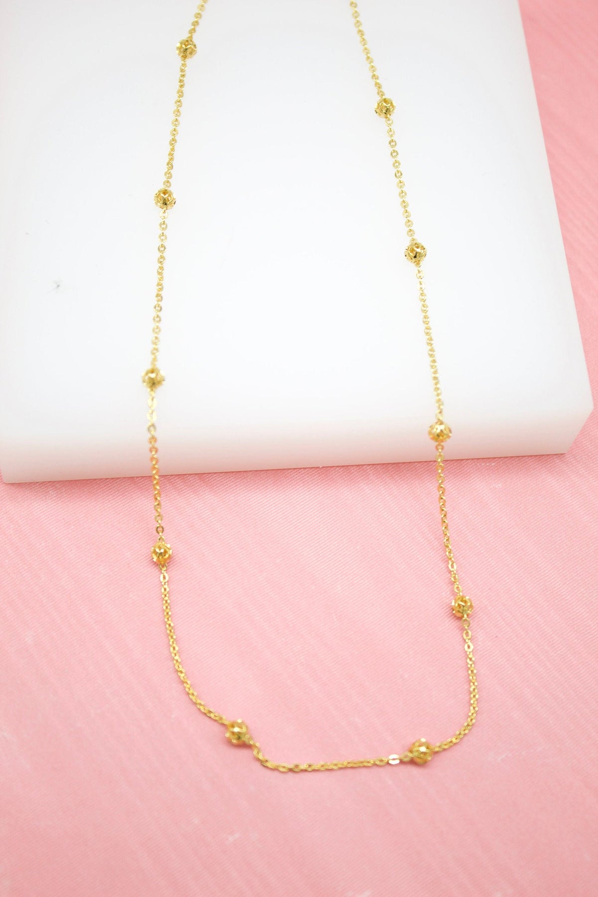 A delicate gold chain necklace with evenly spaced faceted bead stations rests on a white display surface against a pink background.