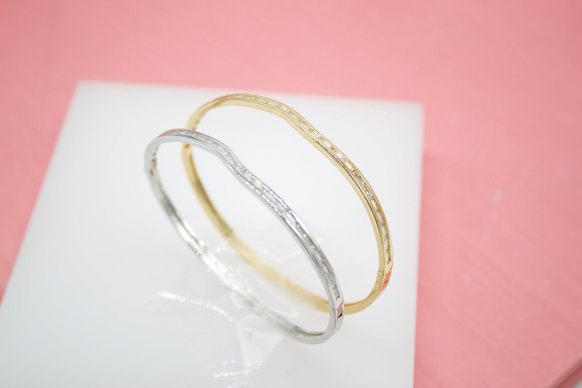 A silver diamond bangle and a gold diamond bangle rest on a white surface against a pink background.