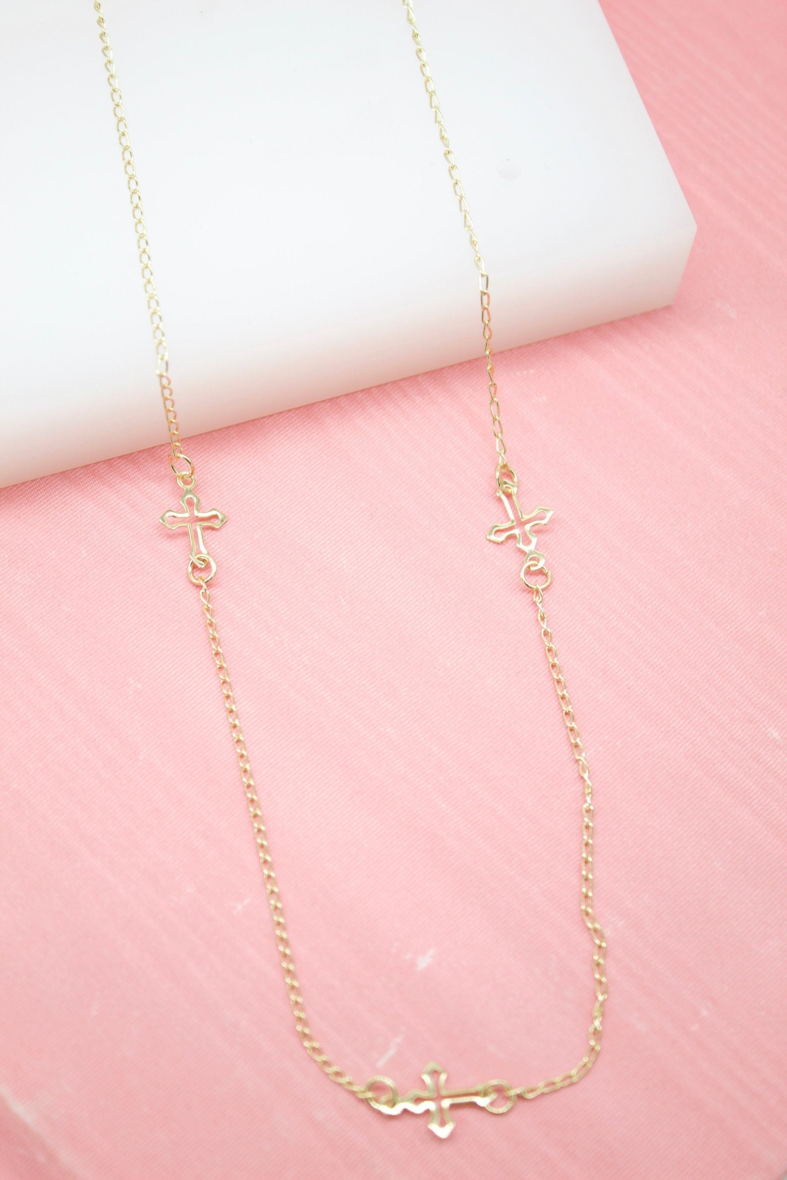 A delicate gold chain necklace features three small cross charms distributed along its length, displayed against a pink background.