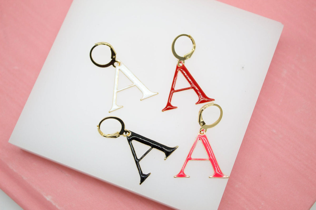 Four colorful enamel letter A leverback earrings rest on a white display surface against a pink background.