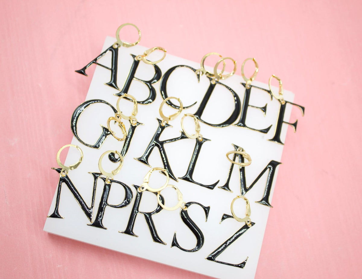 A collection of black enamel alphabet letter earrings with gold hoops rests on a white display block against a pink background.