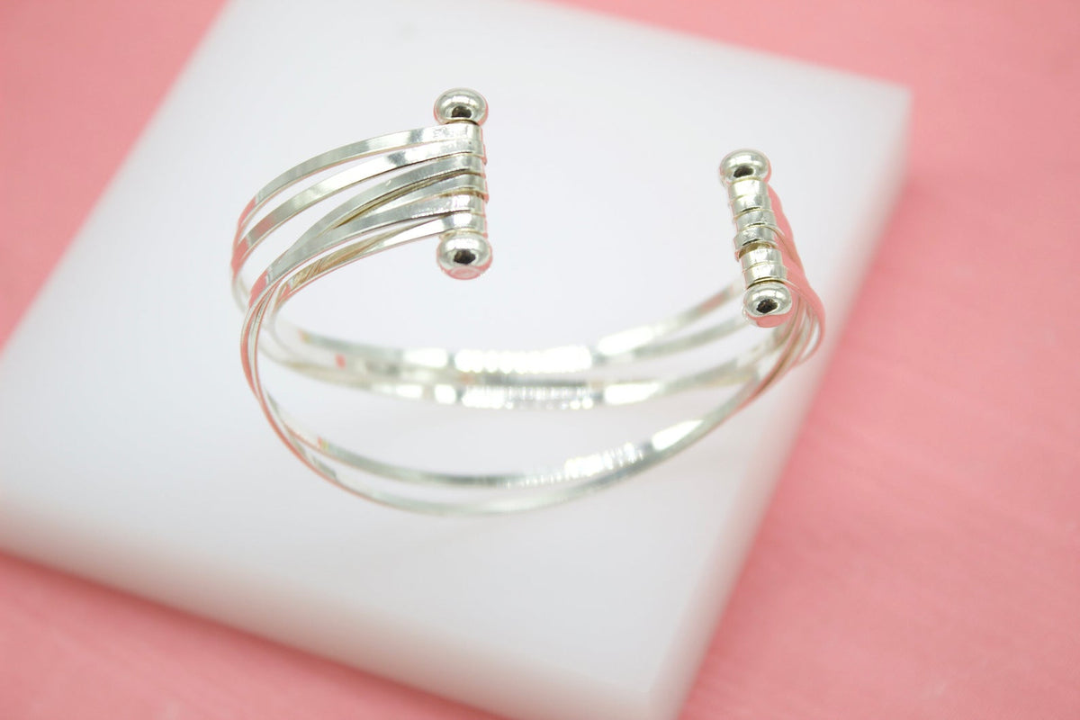 A polished silver multi-band cuff bracelet with ball-end details rests on a white display block against a soft pink background.