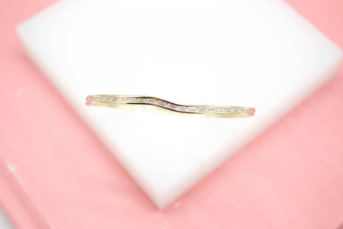 A curved gold band ring set with baguette-cut diamonds rests on a white display block against a soft pink background.