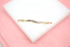 A curved gold band ring set with baguette-cut diamonds rests on a white display block against a soft pink background.